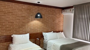 Ein Hotelzimmer mit zwei Betten, einer Ziegelwand, einer hängenden Pendelleuchte, einer Klimaanlage und einem Fenster mit Jalousien.