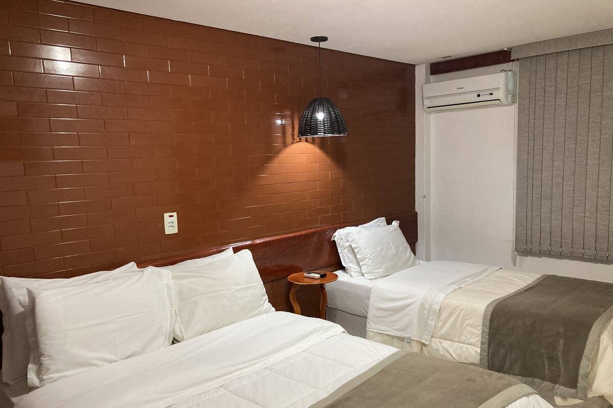 Ein Hotelzimmer mit zwei Betten, einer Ziegelwand, einer Pendelleuchte, einer Klimaanlage und einem Fenster mit Vorhängen.