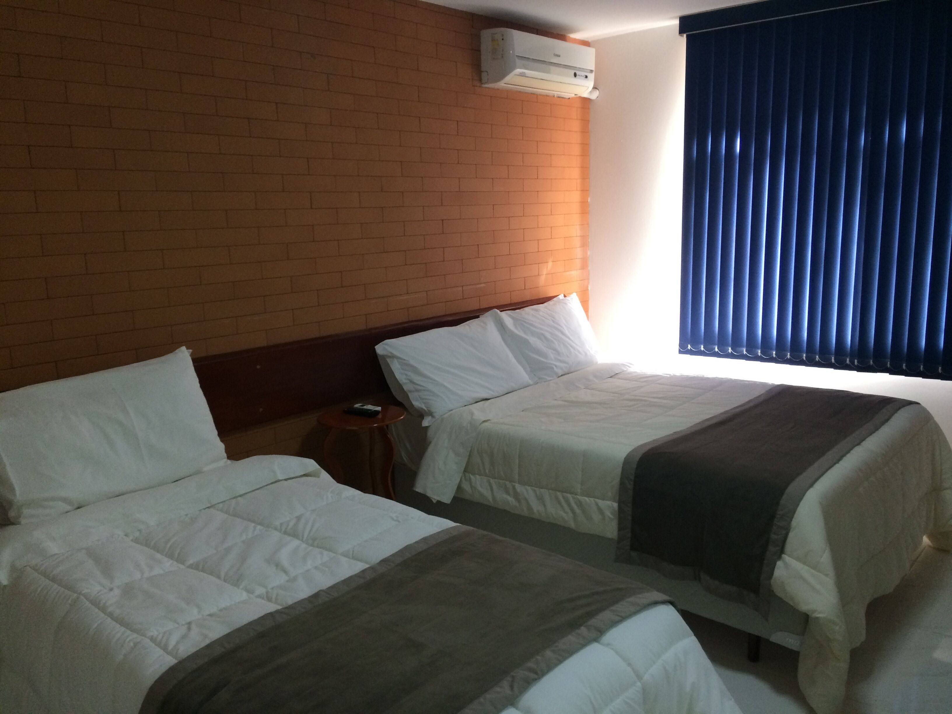 Ein Hotelzimmer mit zwei Betten, einer Ziegelwand, einer Klimaanlage, einem kleinen Tisch und einem Fenster mit blauen Vorhängen.