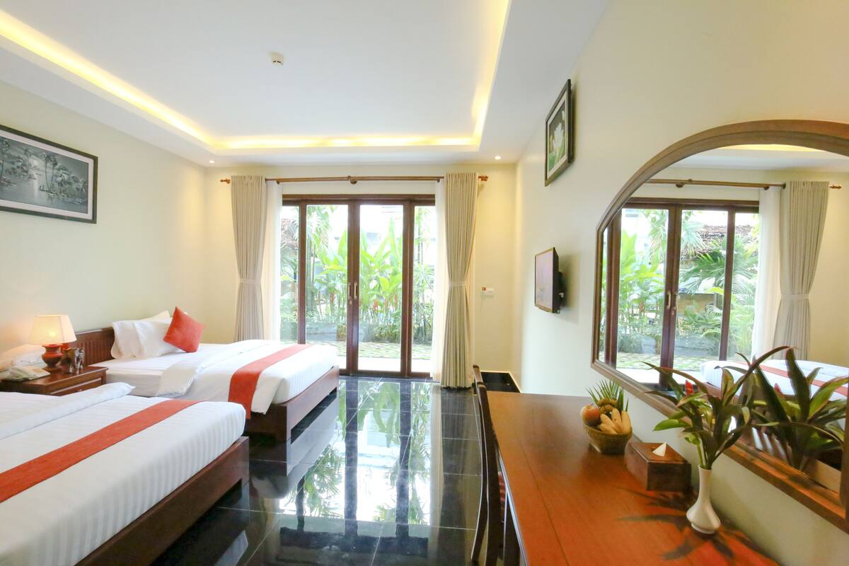 A hotel room with two beds, a wooden desk, a mirror, and a view of greenery through large windows.