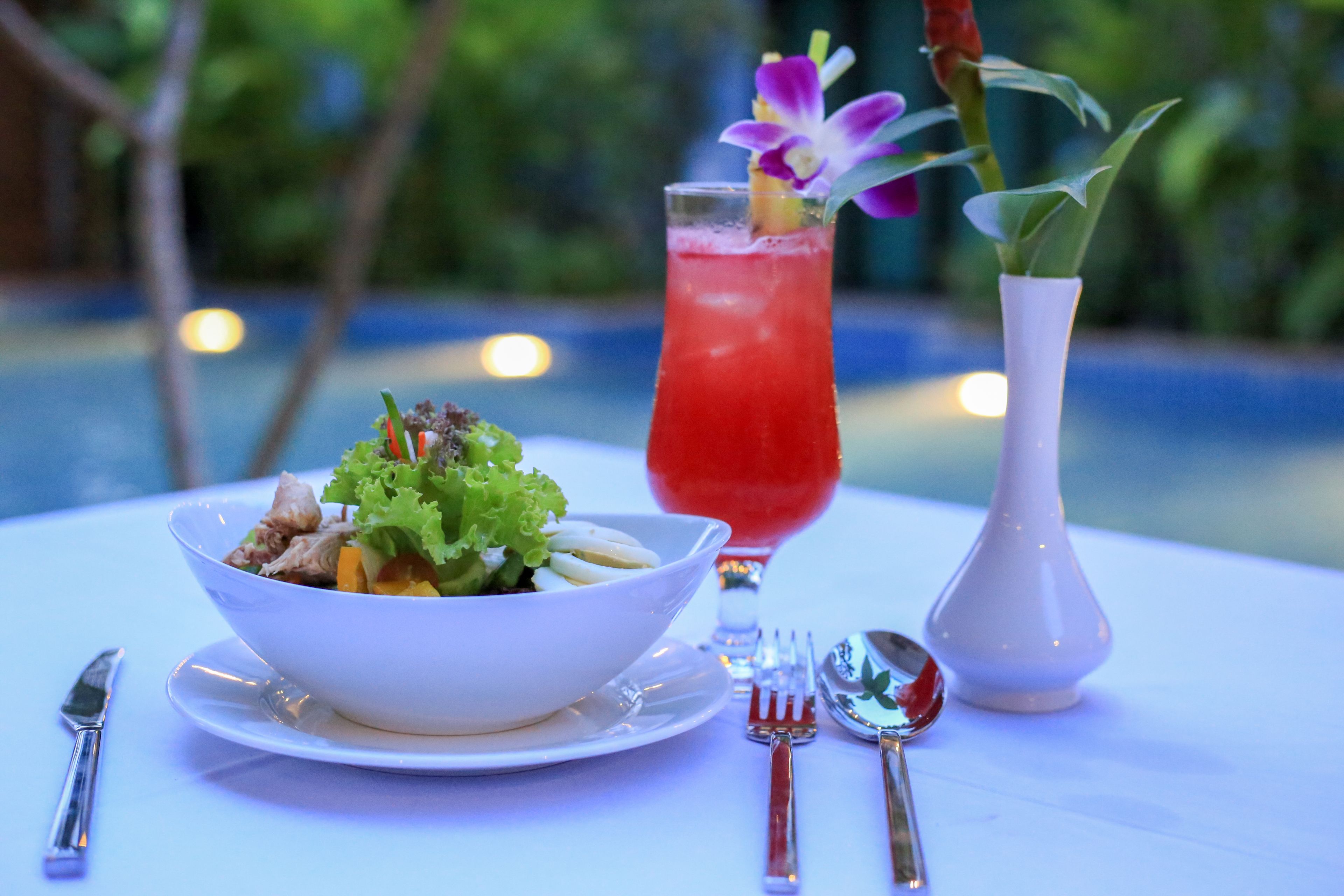 A table with a white bowl of salad, a tall glass of red drink, and a white vase with a flower.