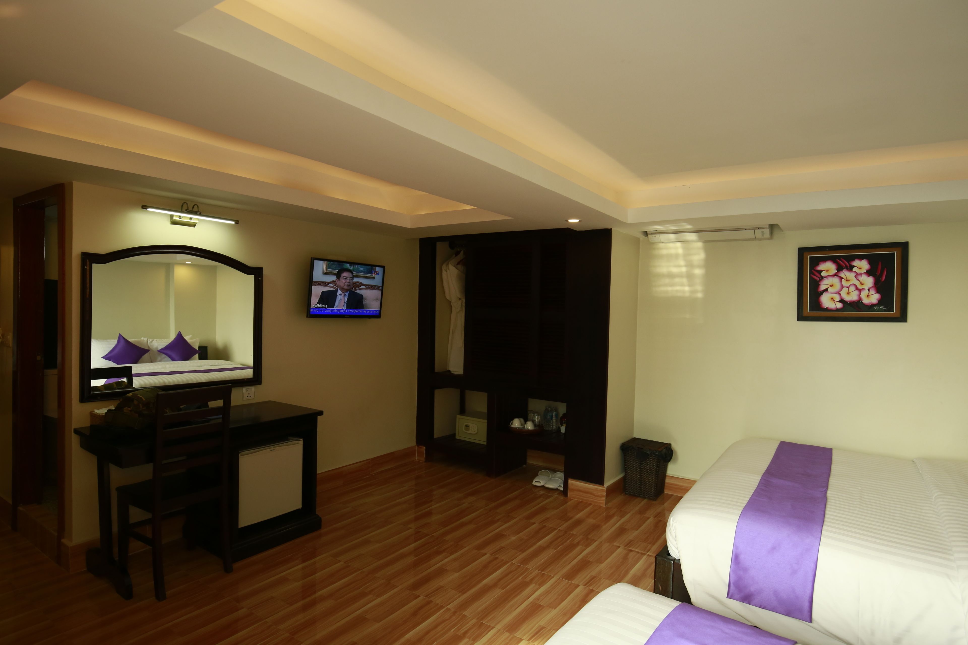 Une chambre d’hôtel avec un plancher en bois, un bureau avec un miroir, une télévision, un lit recouvert d’une courtepointe violette et un tableau encadré accroché au mur.