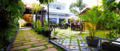 A paved garden pathway leading to outdoor seating areas with tables and chairs, surrounded by potted plants and a building in the background.