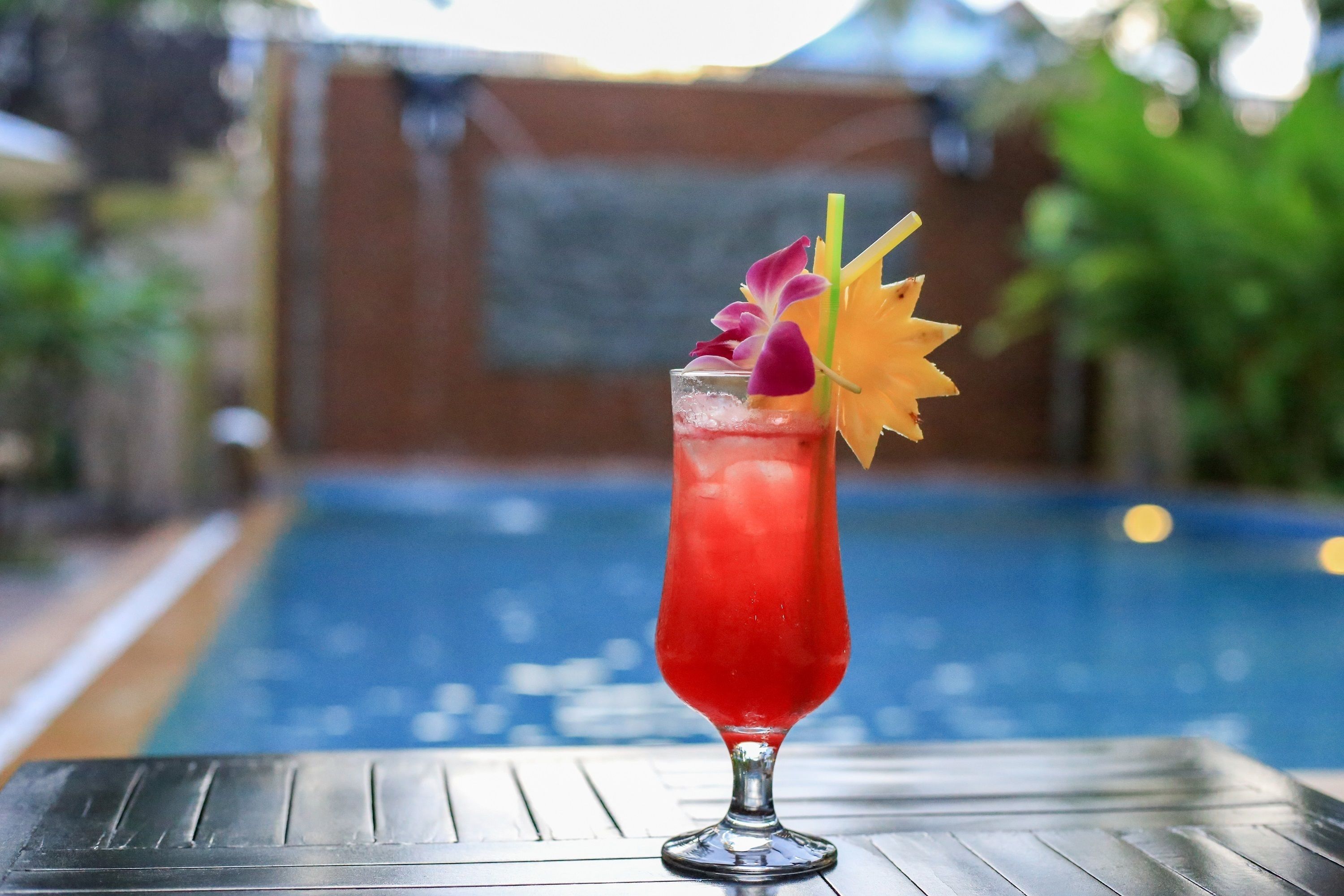 A cocktail in a tall glass with a decorative flower and straw, placed on a table near a swimming pool.