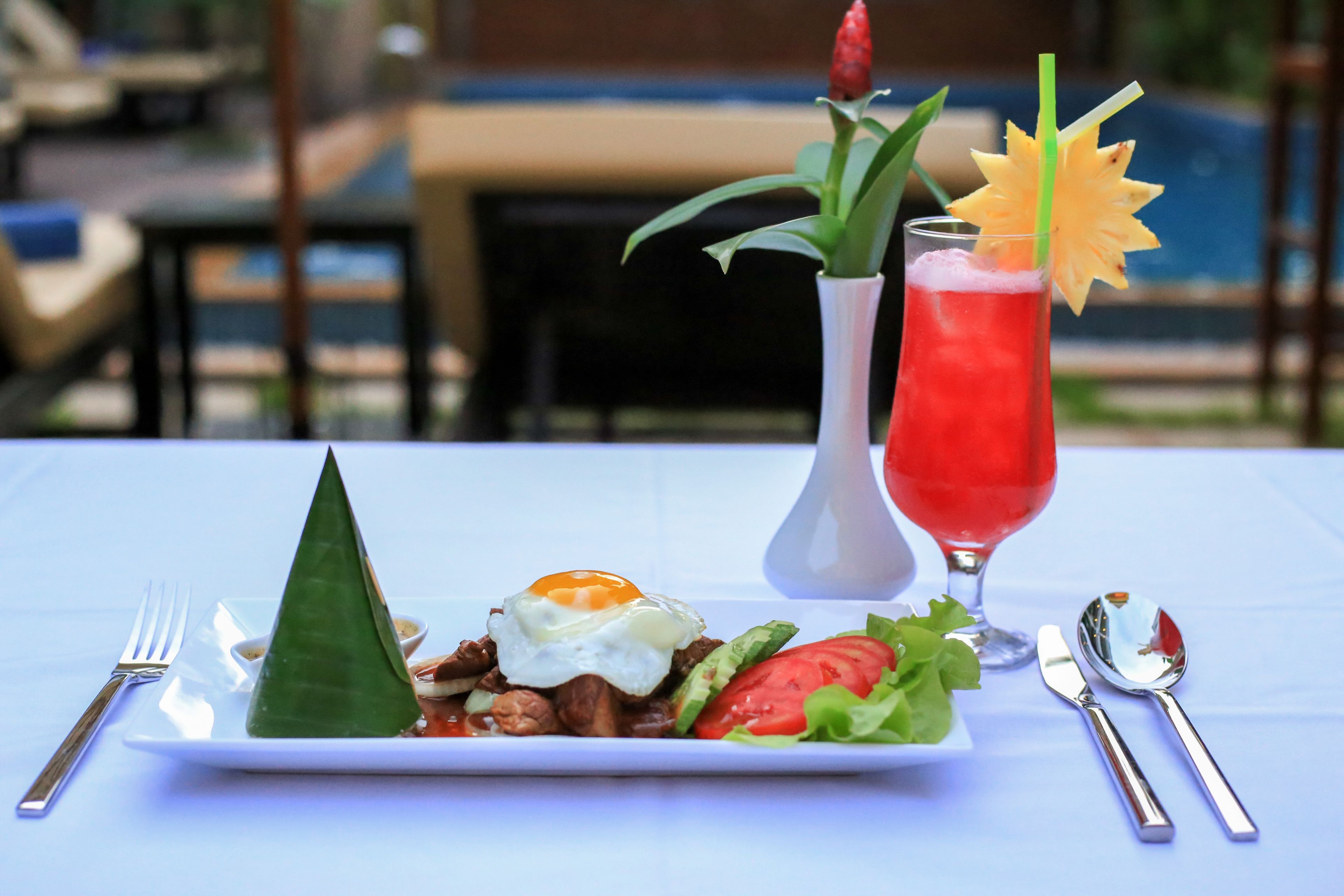 Un plato de comida con un cono verde, un huevo y vegetales, un vaso de bebida roja con una pajita, y un jarrón con una planta.