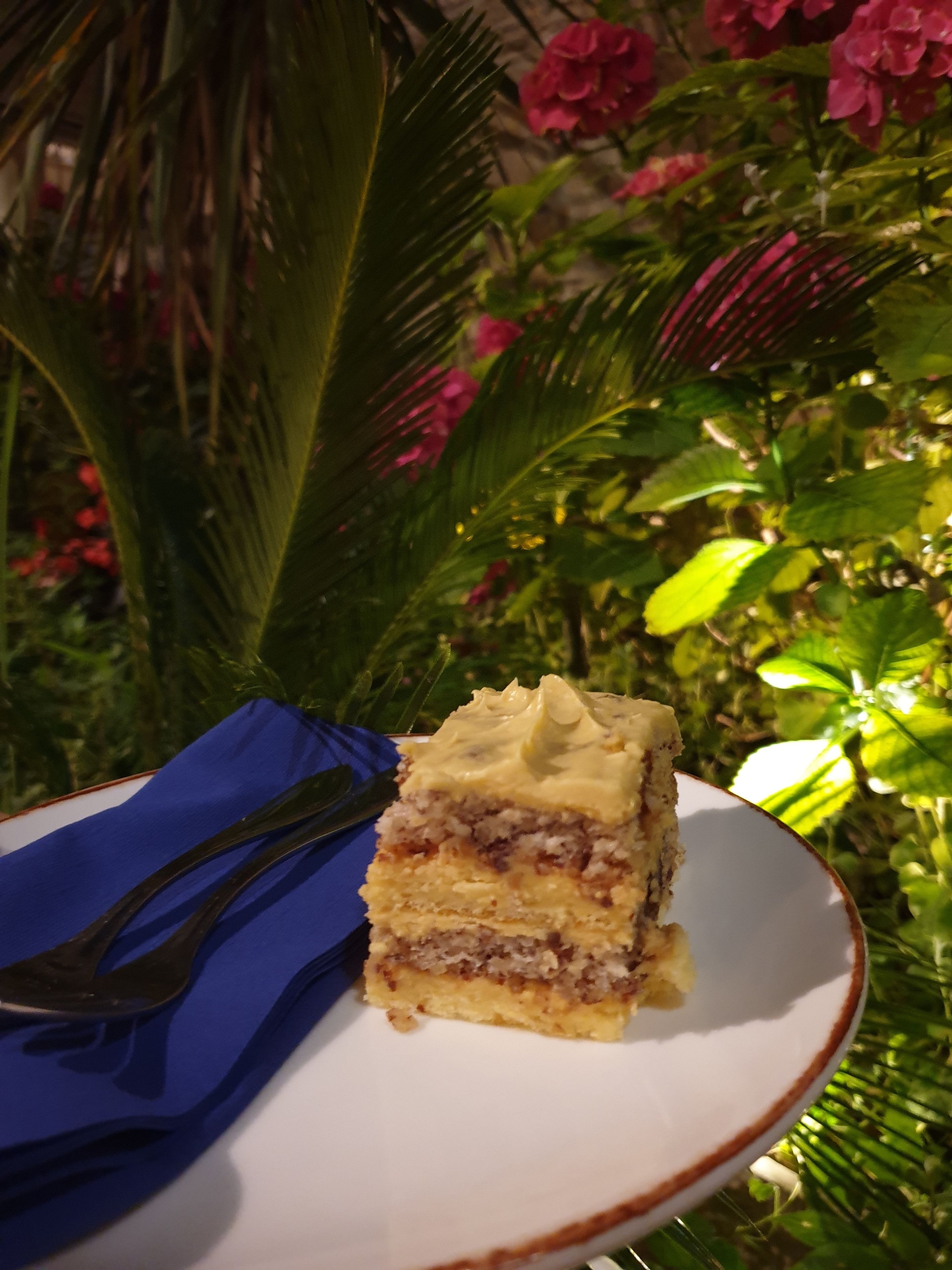 Ein Stück Kuchen auf einem Teller mit einer Gabel, auf einem blauen Serviettenplatz, vor einem Hintergrund aus Grünpflanzen und rosafarbenen Blüten.