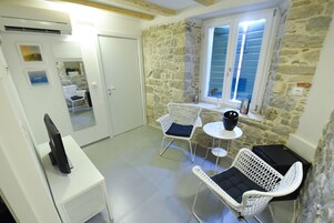 Ein Zimmer mit Stehwänden, einer weißen Tür, einem weißen Stuhl mit Kissen, einem kleinen Tisch und einem Fernseher auf einem Standfuß.