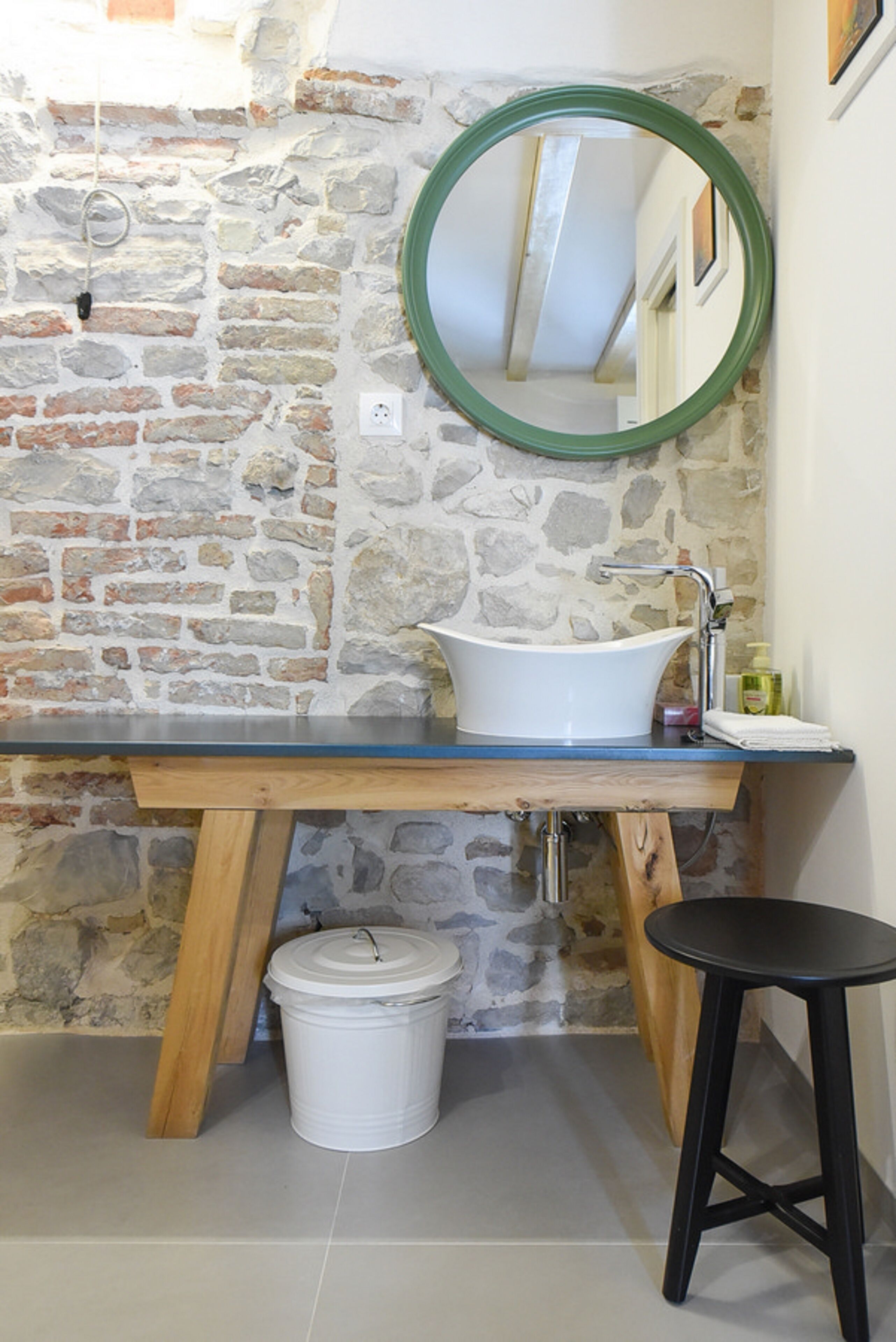 A bathroom with a stone wall, a white sink, a wooden vanity, a black stool, and a white trash bin.