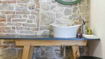 A bathroom with a stone wall, a white sink, a wooden vanity, a black stool, and a white trash bin.