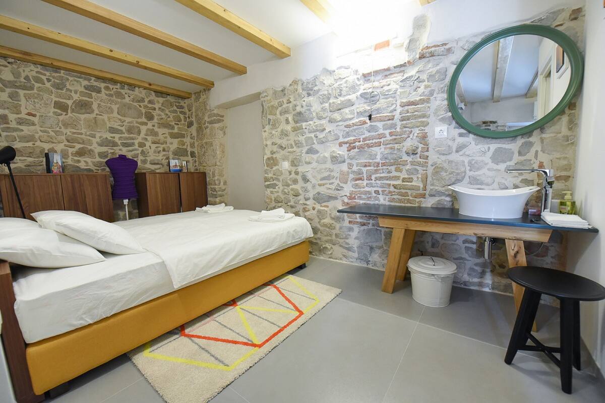 Ein Schlafzimmer mit Bett, Holz-Kopfteil, Steinwand, Badezimmer mit Waschbecken und Spiegel sowie ein kleiner Hocker.