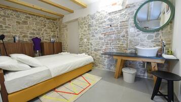 A bedroom with a bed, a wooden headboard, a stone wall, a bathroom with a sink and mirror, and a small stool.