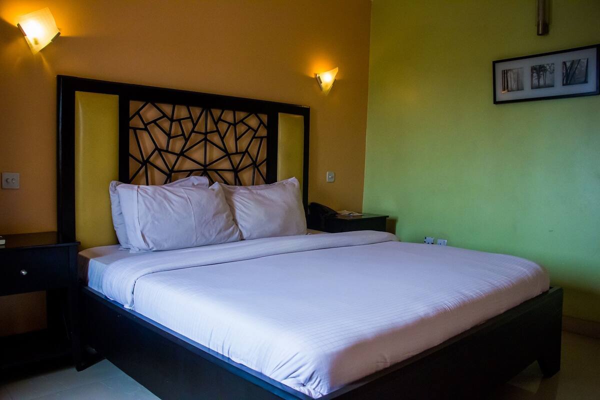 A neatly made bed with white linens, a dark wooden headboard with a geometric pattern, and two wall-mounted light fixtures.