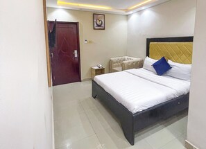 A hotel room with a bed, a small bedside table, a chair, a door, and a wall-mounted picture. - De' Bliss Hotel and Suites (Lagos)