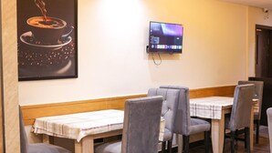 Un coin repas avec des tables en bois et des chaises à revêtement gris, un téléviseur à écran plat fixé au mur et un tableau représentant une tasse de café.