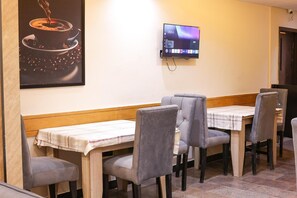 Zona pranzo con tavoli in legno e sedie grigie imbottite, una TV a schermo piatto montata a parete e un quadro con l'immagine di una tazza di caffè.