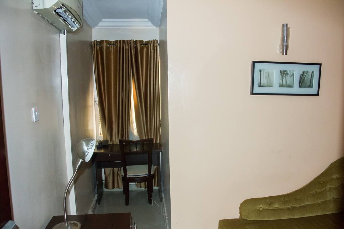 A room with a desk, chair, and a sofa. There is a framed picture on the wall and an air conditioning unit on the wall.
