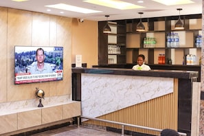 A hotel reception area with a marble counter, a television screen displaying a news broadcast, and shelves stocked with bottled water and snacks.