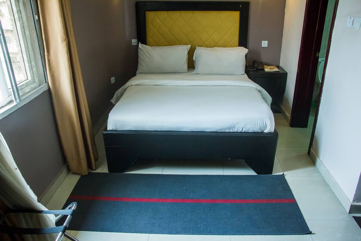 A neatly made bed with white linens and two pillows, a black headboard, and a small bedside table with a phone and a book.