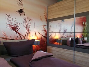 A modern bedroom with a large mirror reflecting a wall mural of tall grasses and a sunset. There are two bedside tables with lamps, a bed with purple bedding, and a small potted plant on the nightstand. - gapart - Apartments mit Küche (Leipzig)