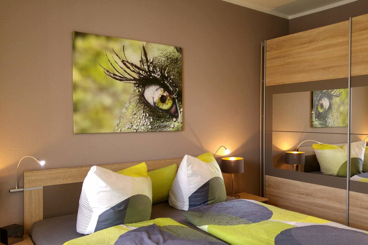 A bedroom with a bed, pillows, a nightstand, a lamp, and a large mirror.