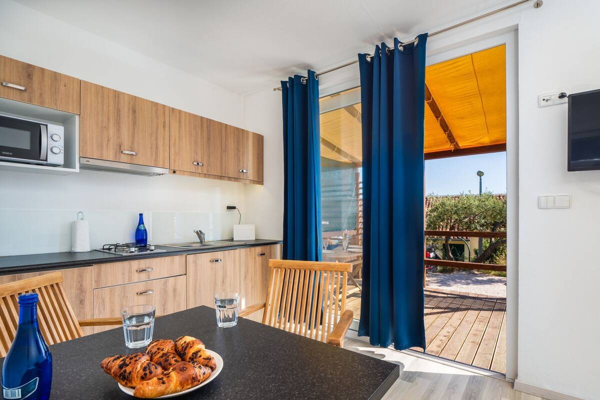 Eine moderne Küche mit Holzschränken, einer schwarzen Arbeitsplatte und einem Essbereich mit Tisch und Stühlen. Auf dem Tisch steht ein Teller mit Croissants und eine Wasserflasche.