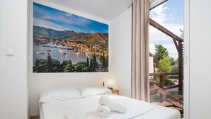 Une chambre d’hôtel avec un lit, un tableau représentant une ville côtière, une fenêtre avec des rideaux et une terrasse en bois à l’extérieur.