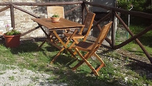 Ein rustikaler Sitzbereich im Freien mit einem Holztisch, Stühlen, einem Blumentopf und einem Steinhaus im Hintergrund.