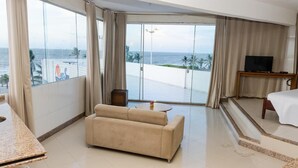 Habitación de hotel con un amplio ventanal corredero que da a un balcón con vistas al océano. Hay un sofá color beige, una mesita redonda y una cama con edredón blanco.