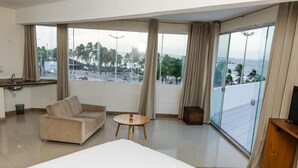 Un cuarto de hotel con una cama grande, un sofá, una mesita, un mueble para televisión y un balcón con vistas a la ciudad y palmeras.