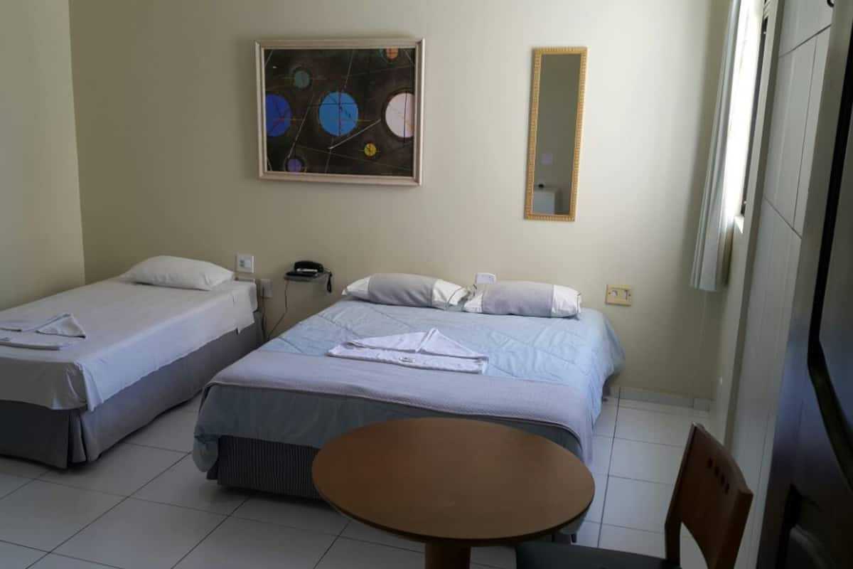 A hotel room with two beds, a small table, a chair, a painting on the wall, and a mirror.