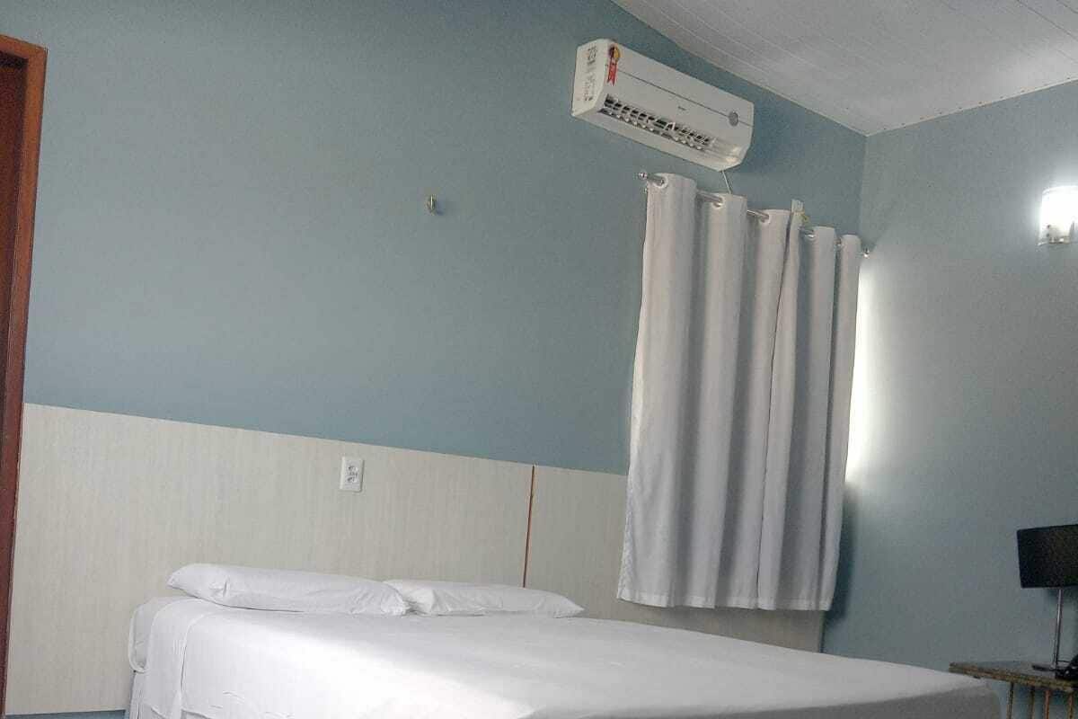A single-bed room with a white bed, a ceiling-mounted air conditioner, a curtain, and a small table with a chair.