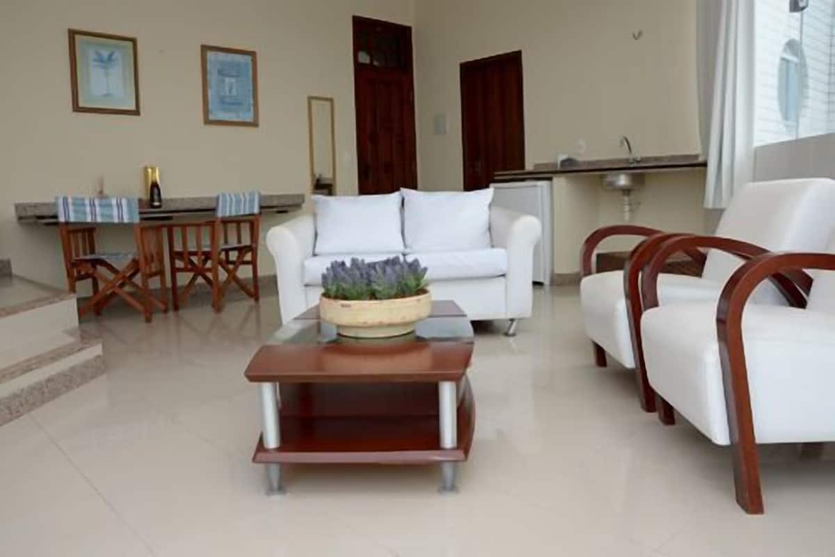 A living room with a white sofa, wooden chairs, a coffee table with a plant, and a kitchen area in the background.