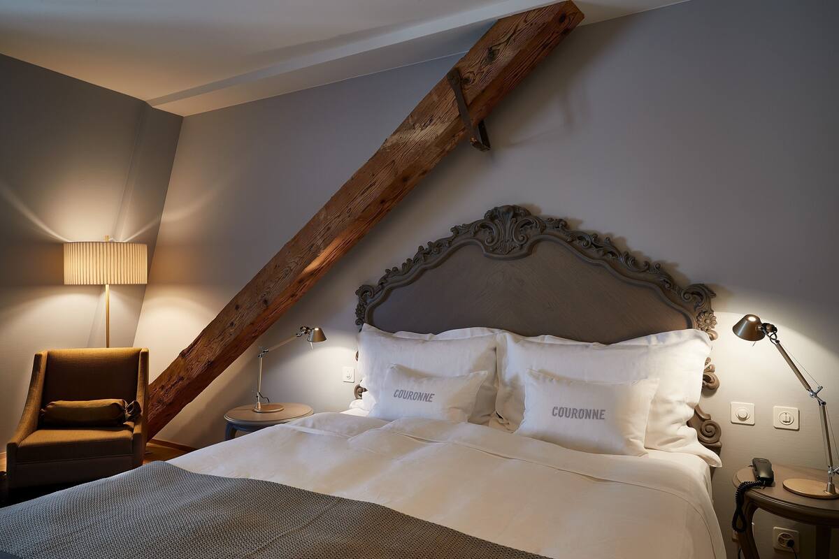 A bedroom with a sloped ceiling, a wooden headboard, a chair, a bedside table with a lamp, and a bed with white bedding and pillows labeled "COURONNE".