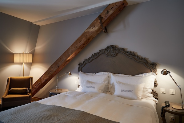 Quarto com teto inclinado, cabeceira de madeira, uma cadeira, mesa de cabeceira com abajur, e cama com roupa de cama e almofadas com a etiqueta "COURONNE".