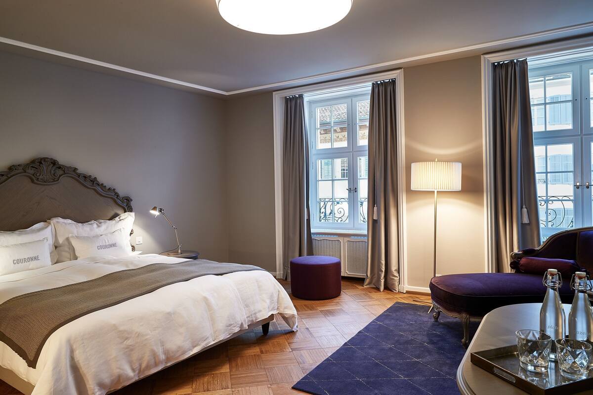 A hotel room with a large bed, a round ottoman, a desk with a lamp, and a view of the outside through a window.