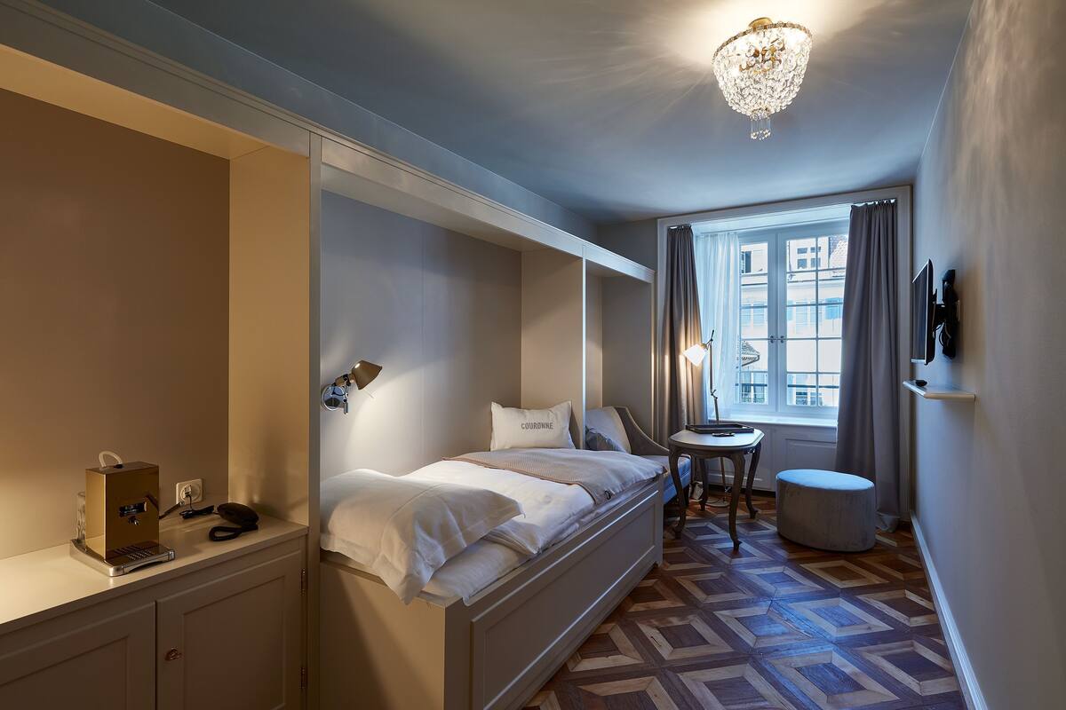 A hotel room with a bed, bedside table, lamp, and a view of the city through a window.
