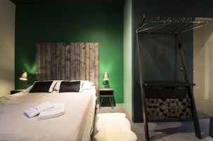 Quarto de hotel com cama, cabeceira de madeira, mesa de cabeceira com luminária, pequena mesa com toalha estampada e um cabideiro.