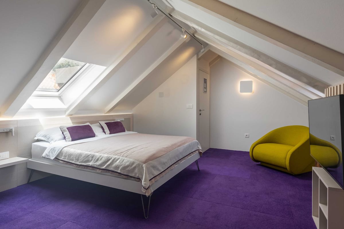 Ein modernes Schlafzimmer mit schrägverdeckter Decke, einem Bett, einem gelben Sessel und einem Dachfenster.
