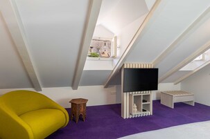 Um quarto com teto inclinado, uma cadeira amarela, um banquinho de madeira e um móvel de entretenimento branco, além de um tapete roxo.
