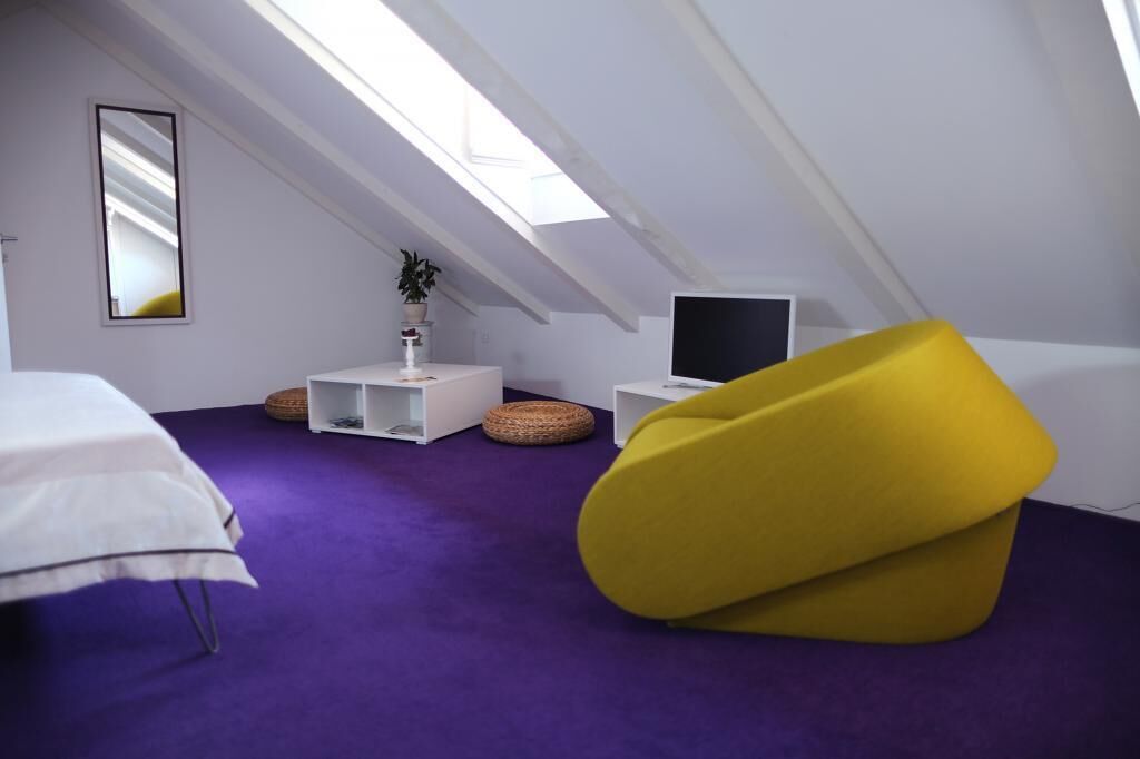 Una habitación moderna y compacta con techo inclinado, una cama, un sillón de mimbre amarillo, una pequeña mesa blanca con una planta y un televisor de pantalla plana.