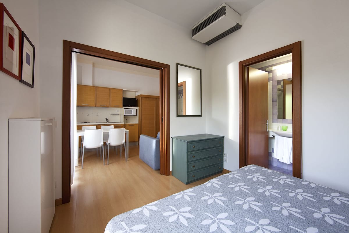 A bedroom with a bed, a dresser, a mirror, and a view into a kitchen area.