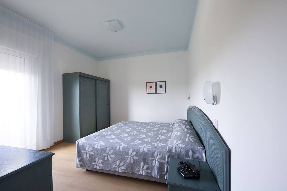 A bedroom with a bed, a wardrobe, a bedside table with a telephone, and a wall-mounted light fixture.
