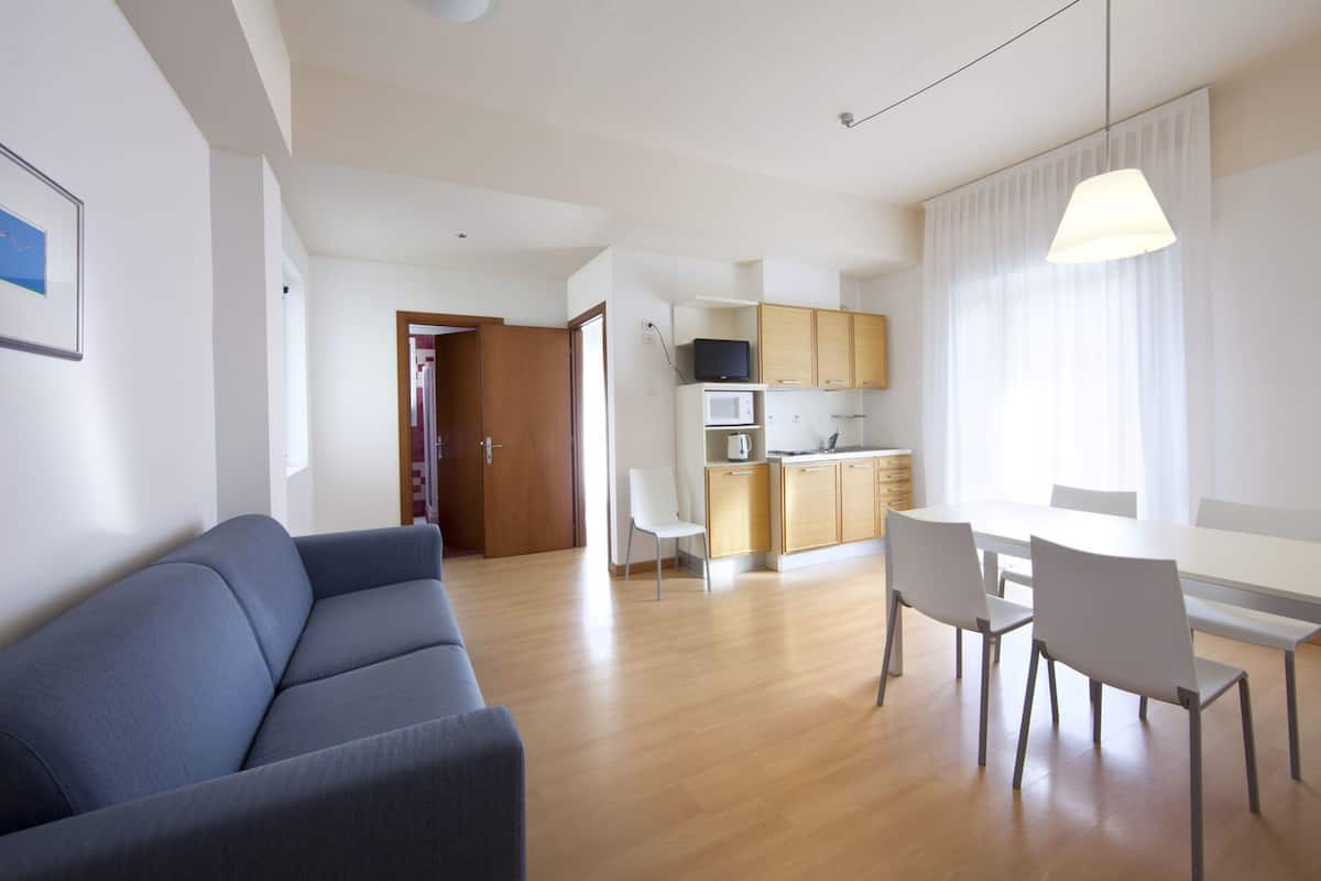 A modern living room with a sofa, a dining area with chairs, a kitchen with cabinets and appliances, and a door leading to another room.