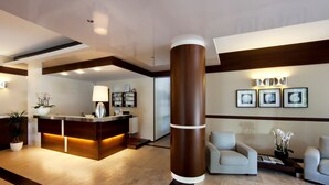 A modern reception area with a wooden reception desk, a clock, framed pictures on the wall, and two sofas. - Aparthotel la Pineta (Jesolo)