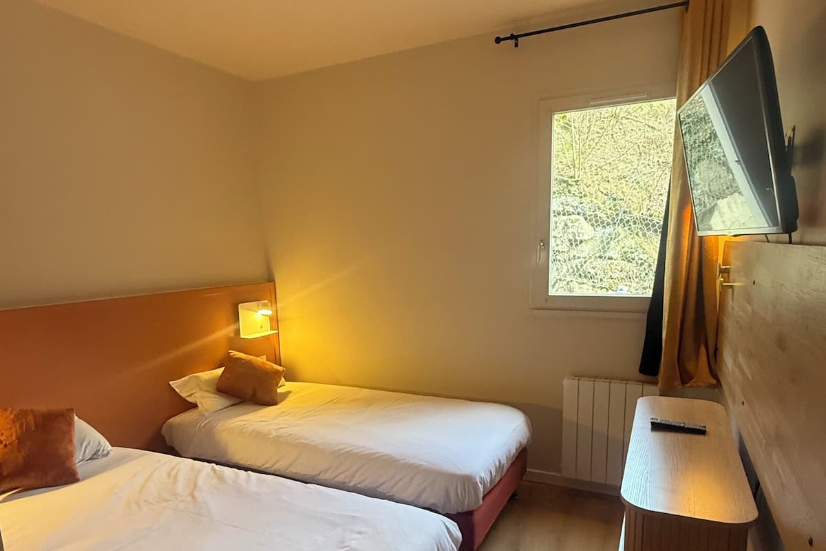 A hotel room with two beds, a desk, a window with curtains, and a radiator.