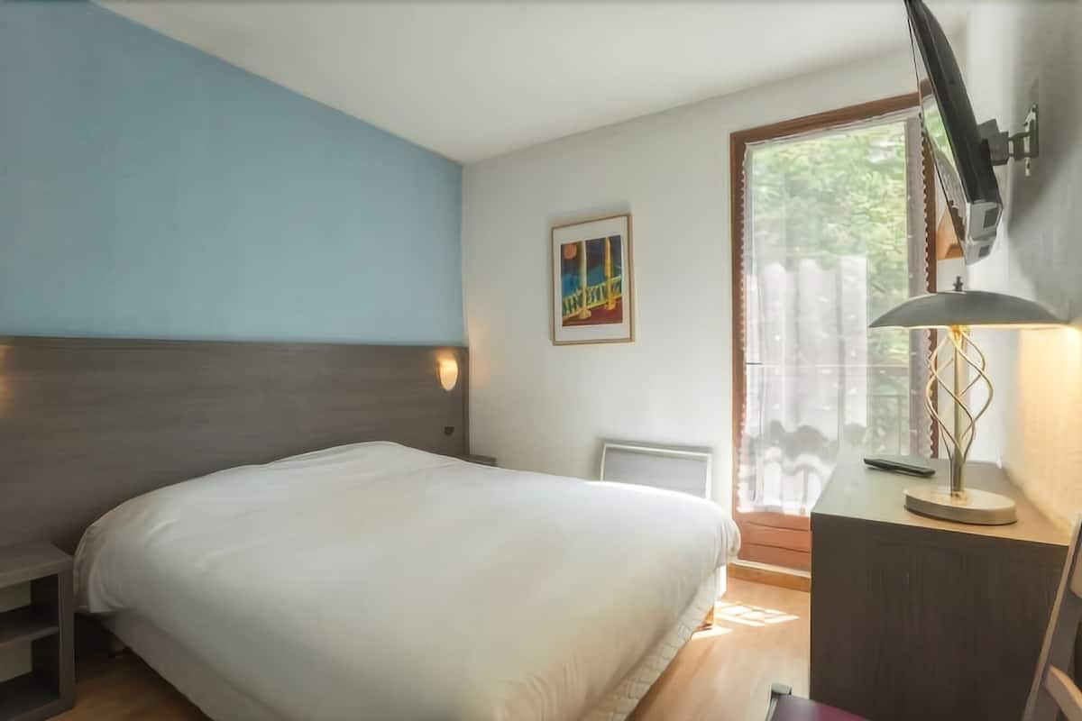A bedroom with a bed, a bedside table, a lamp, a wall-mounted picture, and a window with a view of greenery.