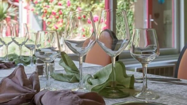 A table set with wine glasses and a metal plate, with a view of a garden with pink flowers through a window. - Hôtel La Cascade (Saint-Genis-les-Ollieres)