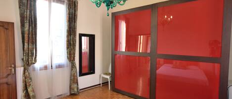 A room with a bed, a mirror, a red sliding door, a chandelier, and a window with curtains.