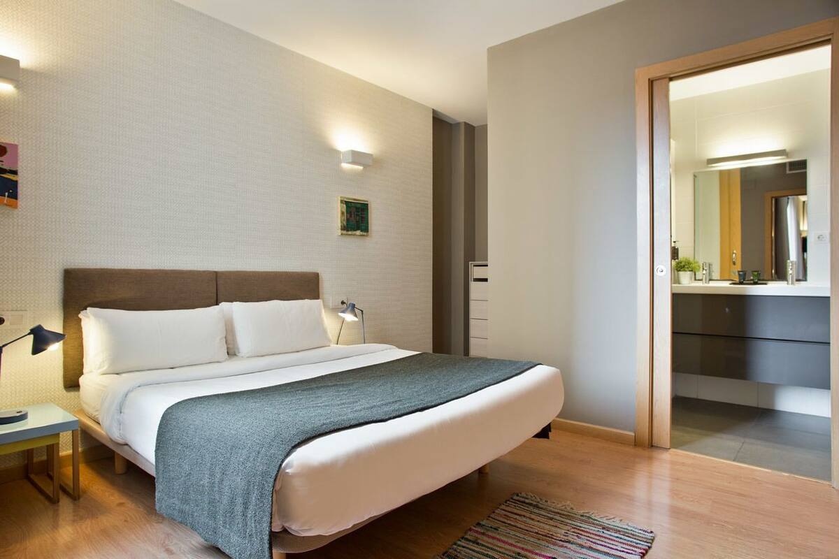 Une chambre d’hôtel moderne, avec un lit soigneusement fait, une table de chevet avec une lampe, un petit tableau accroché au mur et une vue sur la salle de bain par une porte entrouverte.