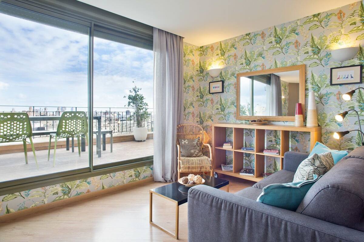 Un salon avec un balcon, un fauteuil en osier, un canapé gris et une bibliothèque.